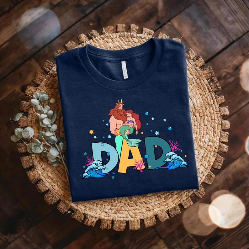 May include: Navy blue t-shirt with a graphic of King Triton from The Little Mermaid holding Ariel. The graphic is surrounded by stars, bubbles, and waves. The text "DAD" is in large, colorful letters below the graphic.