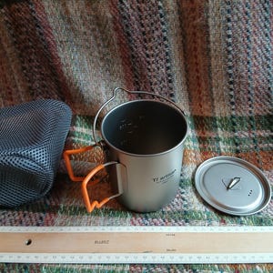 May include: A titanium camping mug with orange handles and a lid, next to a mesh storage bag. The mug has a foldable handle and measurement markings inside. A wooden ruler is at the bottom.