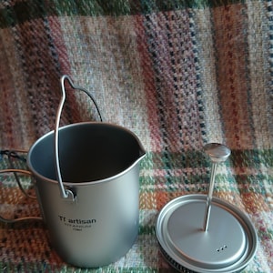 May include: A silver titanium camping coffee pot with a handle and a lid. The pot has the text "Ti artisan TITANIUM 750ml" printed on the side. The lid has a central post with a small circular handle. The pot is on a plaid surface.