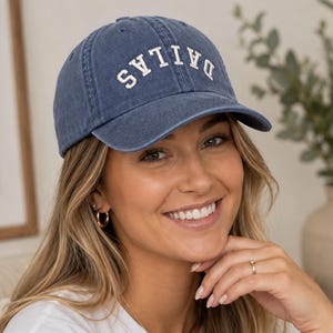 May include: A blue baseball cap with a curved brim and the word DALLAS embroidered in white block letters across the front. The cap features a washed-out denim texture, a structured crown, and a button on top, providing a classic athletic look.