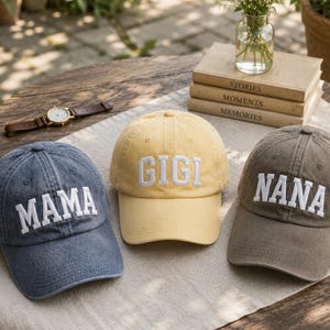 May include: Three baseball caps in blue, yellow, and brown, each featuring white embroidered text on the front. The blue cap displays the word MAMA, the yellow cap displays the word GIGI, and the brown cap displays the word NANA in block letters.
