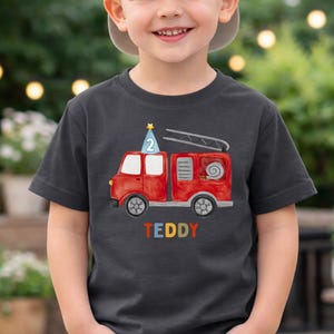 May include: A dark gray short-sleeve t-shirt featuring a red fire truck graphic with a blue party hat displaying the number 2 on top. The name TEDDY is printed in multicolored letters below the fire truck illustration on the front of the shirt.
