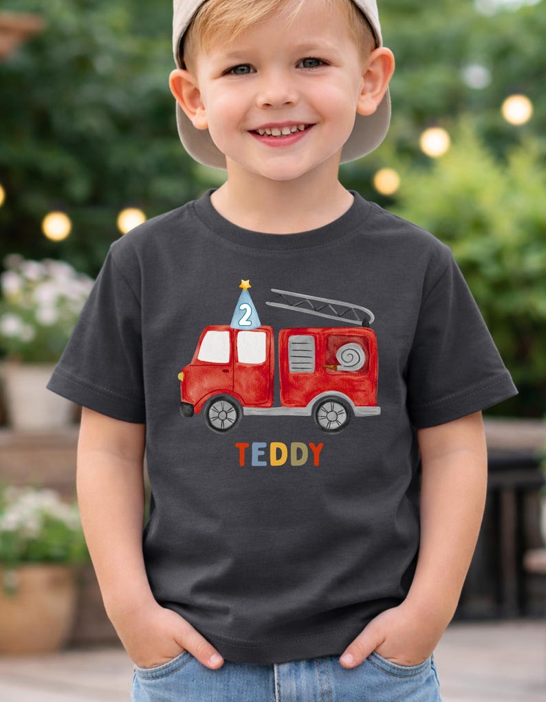 May include: A dark gray short-sleeve t-shirt featuring a red fire truck graphic with a blue party hat displaying the number 2 on top. The name TEDDY is printed in multicolored letters below the fire truck illustration on the front of the shirt.