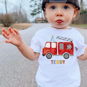 May include: A white short-sleeved t-shirt featuring a watercolor-style red fire truck graphic. The truck has a blue party hat with the number 2 on top and a ladder. The name TEDDY is printed below the truck in multicolored block letters.