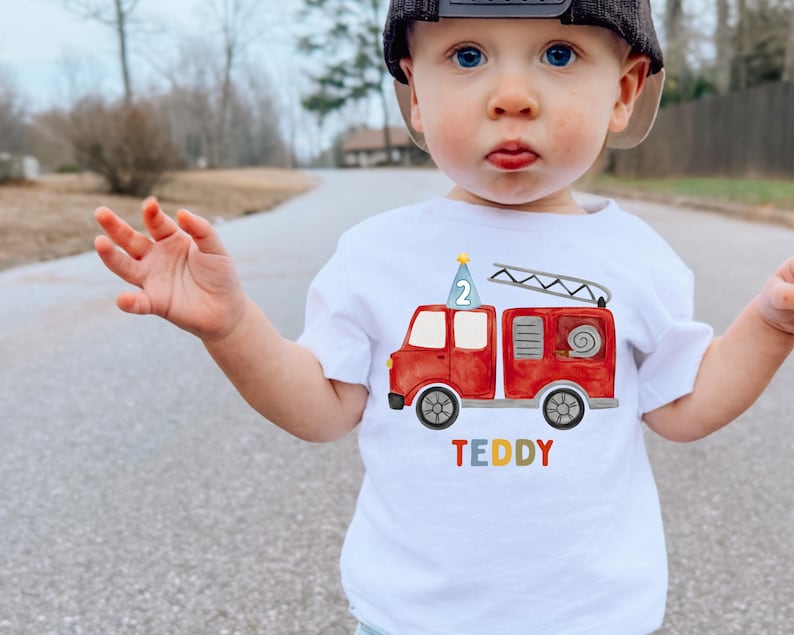 May include: A white short-sleeved t-shirt featuring a watercolor-style red fire truck graphic. The truck has a blue party hat with the number 2 on top and a ladder. The name TEDDY is printed below the truck in multicolored block letters.