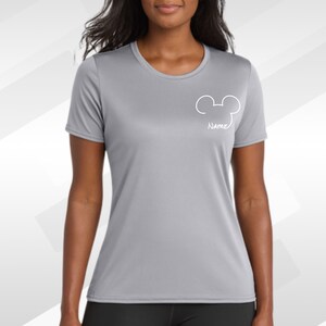 May include: A light gray athletic t-shirt with a white Mickey Mouse head outline and the word "Name" below it. The shirt has a crew neck and short sleeves. The background is a gradient of gray and white.