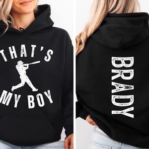 K&ouml;nnte beinhalten: Schwarzer Hoodie mit wei&szlig;em Text, der "That's My Boy" sagt, und einer Baseballspieler-Grafik auf der Vorderseite. Die R&uuml;ckseite des Hoodies hat wei&szlig;en Text, der "Brady" sagt.