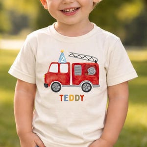 May include: A cream-colored short-sleeve t-shirt featuring a red fire truck graphic with a blue party hat on top displaying the number 2. Below the fire truck, the name TEDDY is printed in multicolored letters. The shirt has a crew neckline.