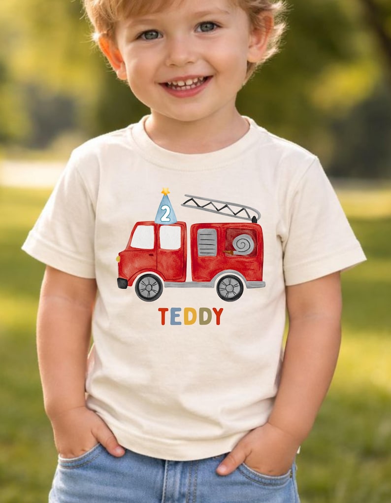 May include: A cream-colored short-sleeve t-shirt featuring a red fire truck graphic with a blue party hat on top displaying the number 2. Below the fire truck, the name TEDDY is printed in multicolored letters. The shirt has a crew neckline.