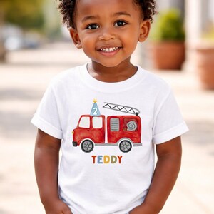 May include: A white short-sleeved t-shirt featuring a watercolor-style red fire truck graphic. The truck has a blue party hat with the number 2 on top and a ladder. Below the truck, the name TEDDY is printed in multicolored block letters.