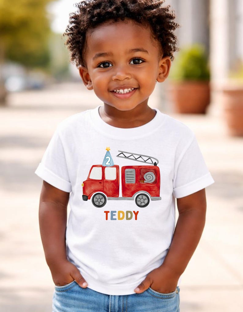 May include: A white short-sleeved t-shirt featuring a watercolor-style red fire truck graphic. The truck has a blue party hat with the number 2 on top and a ladder. Below the truck, the name TEDDY is printed in multicolored block letters.