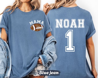 May include: Blue jean-colored t-shirt with the word "MAMA" above a football graphic on the front. The back of the shirt displays the name "NOAH" and the number "1" in white block lettering. The shirt has rolled-up sleeves.