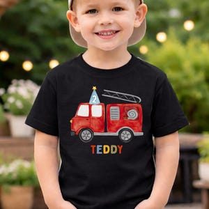 May include: A black short-sleeved t-shirt featuring a watercolor-style red fire truck graphic. The truck has a blue party hat with the number 2 on top and a ladder. The name TEDDY is printed in colorful block letters below the fire truck.