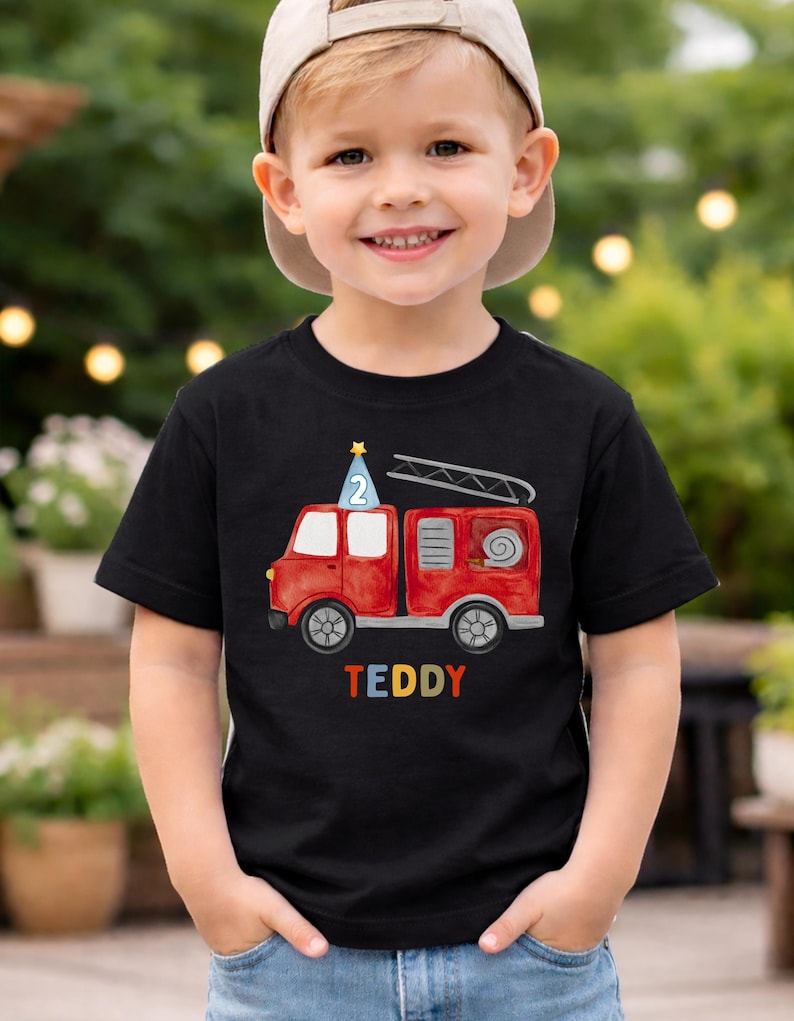 May include: A black short-sleeved t-shirt featuring a watercolor-style red fire truck graphic. The truck has a blue party hat with the number 2 on top and a ladder. The name TEDDY is printed in colorful block letters below the fire truck.