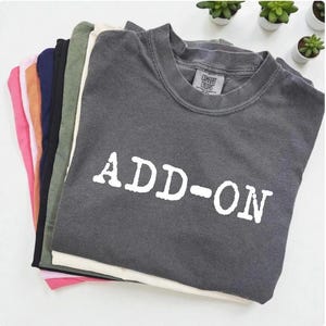 May include: A stack of various colored t-shirts, including pink, orange, navy, olive green, and charcoal gray. The top shirt is charcoal gray with the white text "ADD-ON". The shirts are folded and stacked neatly.