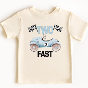 Może przedstawiać: Kremowy t-shirt z kr&oacute;tkim rękawem z grafiką jasnoniebieskiego zabytkowego samochodu wyścigowego z numerem 2 na boku. Nad samochodem znajduje się napis TWO w kolorze jasnoniebieskim, otoczony dwiema szachownicami. Poniżej znajduje się napis FAST.