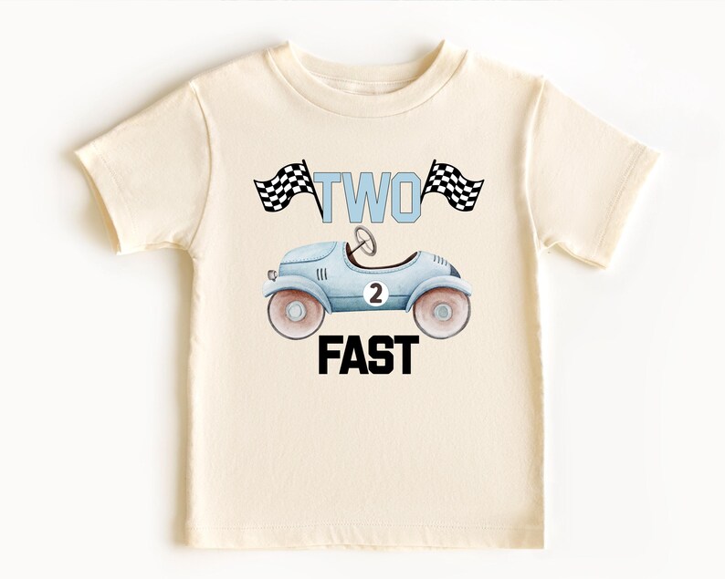 Może przedstawiać: Kremowy t-shirt z kr&oacute;tkim rękawem z grafiką jasnoniebieskiego zabytkowego samochodu wyścigowego z numerem 2 na boku. Nad samochodem znajduje się napis TWO w kolorze jasnoniebieskim, otoczony dwiema szachownicami. Poniżej znajduje się napis FAST.