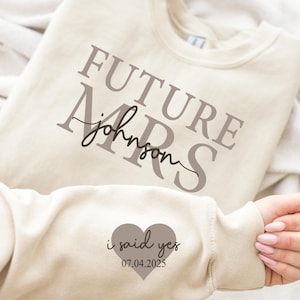 May include: Beige sweatshirt with the text "FUTURE MRS johnson" in brown lettering. A heart with the text "i said yes 07.04.2025" is on the sleeve.