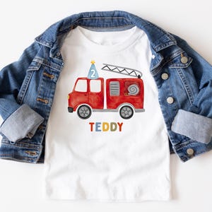 May include: A white t-shirt featuring a watercolor illustration of a red fire truck with a blue party hat displaying the number 2 on top, and the name TEDDY printed in multicolored letters below, layered under a blue denim jacket.