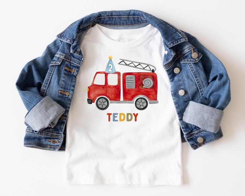 May include: A white t-shirt featuring a watercolor illustration of a red fire truck with a blue party hat displaying the number 2 on top, and the name TEDDY printed in multicolored letters below, layered under a blue denim jacket.