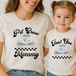 Może przedstawiać: Dwie koszulki w kolorze kremowym z grafiką jasnoniebieskiego samochodu wyścigowego w stylu vintage z numerem jeden, flagami w szachownicę i czarną szachownicą. Koszulka dla dorosłych ma napis Pit Crew Mommy, a dziecięca Fast One.