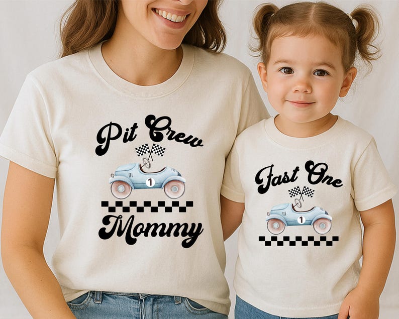 Może przedstawiać: Dwie koszulki w kolorze kremowym z grafiką jasnoniebieskiego samochodu wyścigowego w stylu vintage z numerem jeden, flagami w szachownicę i czarną szachownicą. Koszulka dla dorosłych ma napis Pit Crew Mommy, a dziecięca Fast One.