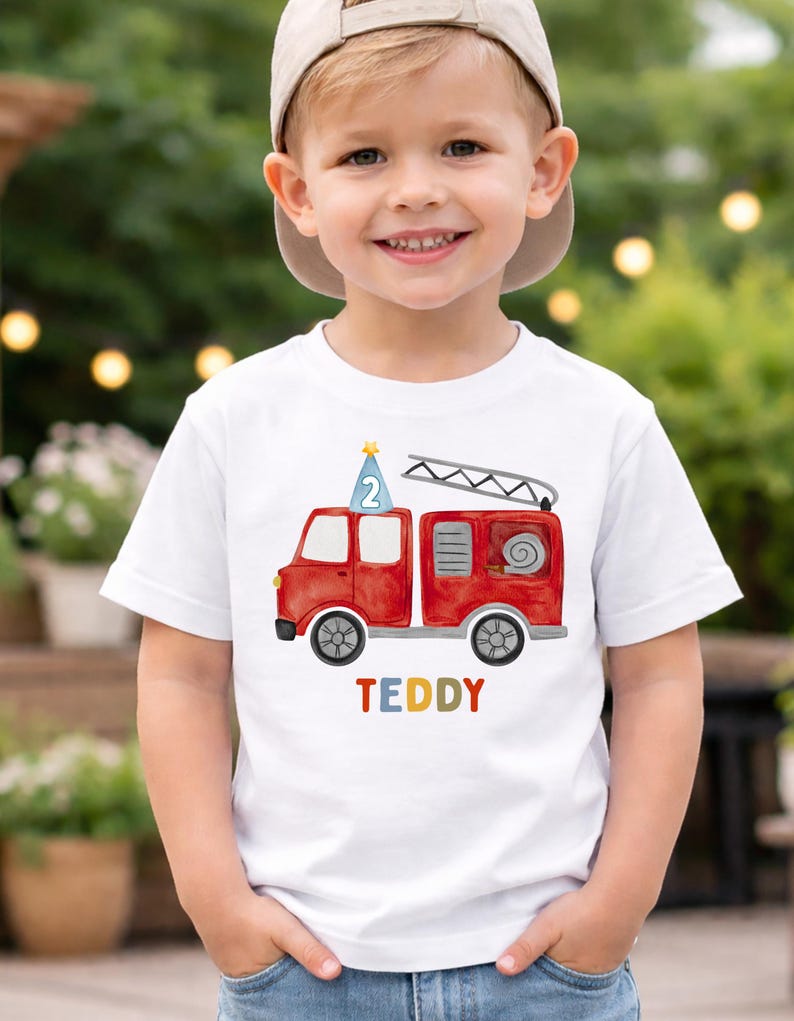 May include: A white t-shirt featuring a red fire truck design with a ladder and the name "TEDDY" in colorful letters. The fire truck has a blue party hat with the number "2" on top. The shirt is worn by a young child.