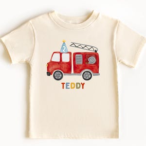 May include: A cream-colored short-sleeve t-shirt featuring a watercolor illustration of a red fire truck with a blue party hat displaying the number 2 on top. The name TEDDY is printed below the truck in multicolored block letters.