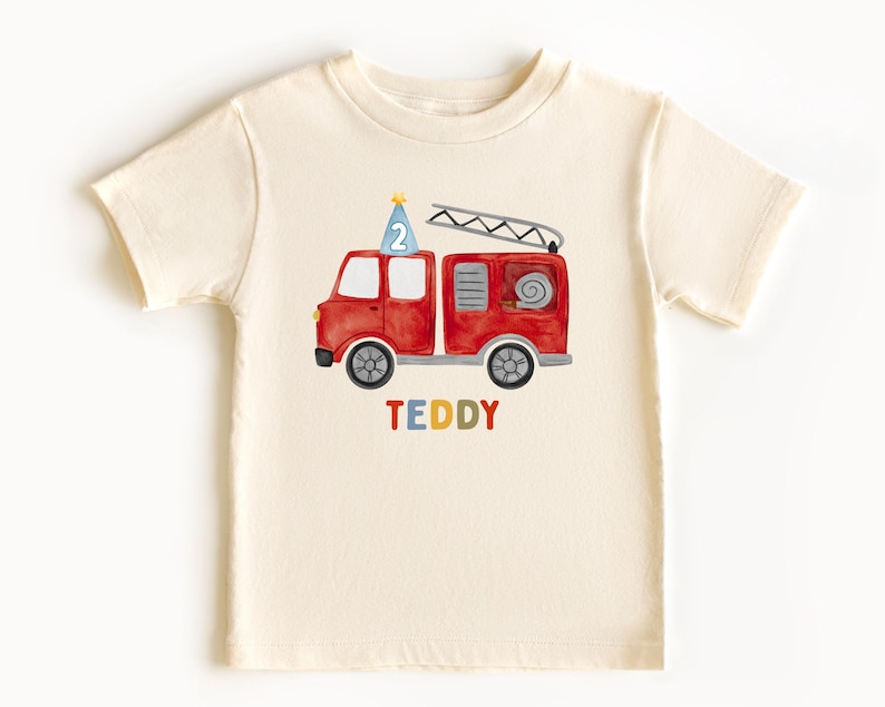 May include: A cream-colored short-sleeve t-shirt featuring a watercolor illustration of a red fire truck with a blue party hat displaying the number 2 on top. The name TEDDY is printed below the truck in multicolored block letters.