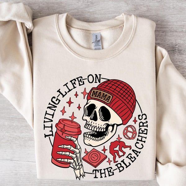 May include: A cream-coloured sweatshirt with a graphic design of a skull wearing a red beanie with the word "MAMA" on it. The skull is holding a red coffee cup and the text "LIVING LIFE ON THE BLEACHERS" is around the skull.