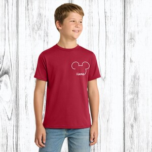 May include: A maroon t-shirt with a white Mickey Mouse head outline and the word "Name" below it. The shirt is short-sleeved and worn by a person. The background is a white wooden wall.