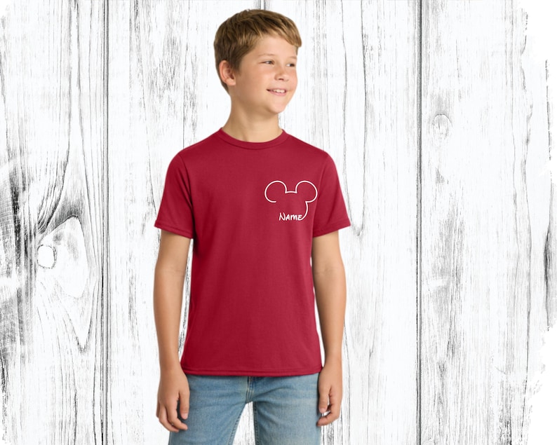 May include: A maroon t-shirt with a white Mickey Mouse head outline and the word "Name" below it. The shirt is short-sleeved and worn by a person. The background is a white wooden wall.