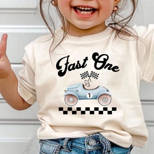 Może przedstawiać: Kremowy t-shirt z kr&oacute;tkim rękawem z czarnym napisem Fast One nad grafiką jasnoniebieskiego zabytkowego samochodu wyścigowego z numerem jeden, dwiema flagami w szachownicę i czarno-białą szachownicą u dołu.