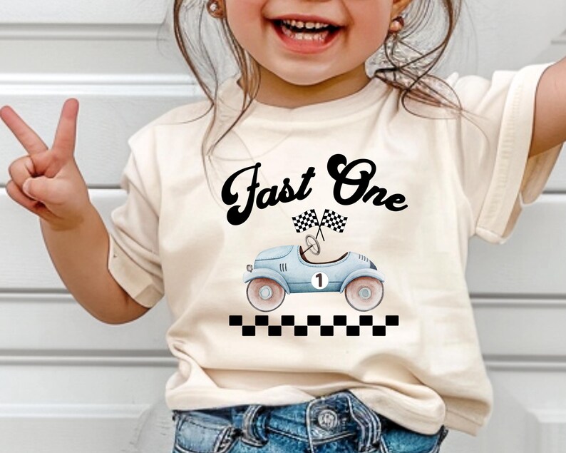 Może przedstawiać: Kremowy t-shirt z kr&oacute;tkim rękawem z czarnym napisem Fast One nad grafiką jasnoniebieskiego zabytkowego samochodu wyścigowego z numerem jeden, dwiema flagami w szachownicę i czarno-białą szachownicą u dołu.