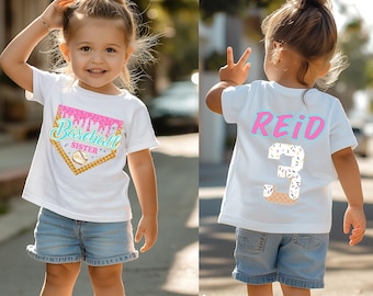 May include: A white short-sleeve t-shirt featuring a graphic of a baseball home plate shaped like an ice cream cone with the text Baseball SISTER. The back displays the name REID in pink and the number 3 designed with colorful sprinkles.