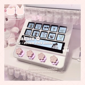 May include: A white stream deck with a grid of small screens displaying cartoon images. The deck has four pink flower-shaped knobs. Text on the deck reads "sakura" and "set of 4 - stream deck knobs".