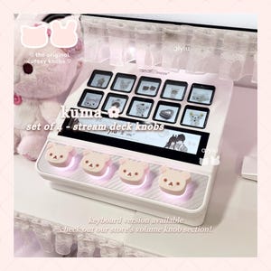 May include: A white stream deck device featuring a set of four light beige bear-shaped knobs with printed facial features. The device includes a screen with multiple icons and text that reads 'kuma set of 4 - stream deck knobs' and 'keyboard version available'.