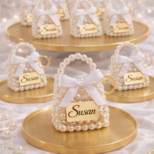 May include: Miniature pearl-covered purse favours with white bows and gold handles, each bearing a gold tag with the name "Susan." The purses sit on gold trays, creating an elegant display.