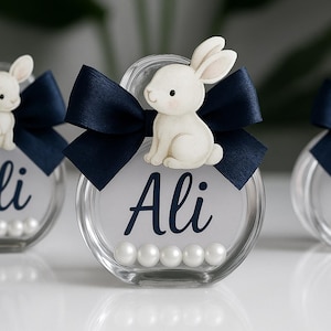 May include: Three clear glass containers, each with a white bunny figurine, a navy blue bow, and the name "Ali" in navy script. The containers are filled with white pearls.