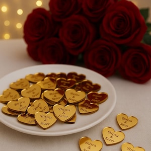 Könnte beinhalten: Goldene herzförmige Gastgeschenke mit den Namen "Büsra & Sezer" sind auf einem weißen Teller zu sehen. Ein Strauß roter Rosen befindet sich im Hintergrund. Die Gastgeschenke sind verstreut.