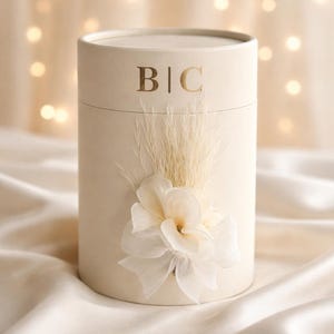 Könnte beinhalten: Eine zylindrische, cremefarbene Geschenkbox mit goldenen Buchstaben "B|C". Die Box ist mit einem Cluster aus getrockneten Gräsern und einer zarten, cremefarbenen Stoffblume verziert. Der Hintergrund ist ein weiches Bokeh aus Lichtern.