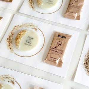 May include: A white and gold round favor with the text "Lorin & Veli 18.01.2025" on a white card with a brown and white illustration of wheat stalks. A brown packet of instant coffee with the text "KURUKAHVECİ MEHMET EFENDİ" is also on the card.