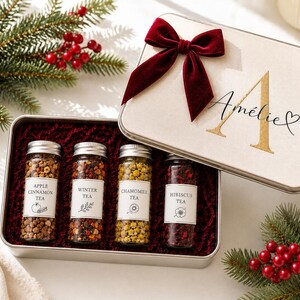 Pode incluir: Uma caixa de presente retangular de metal com um laço de veludo vermelho escuro e o nome Amélie impresso em dourado. Dentro, quatro frascos de vidro com chá a granel rotulados como Apple Cinnamon, Winter, Chamomile e Hibiscus sobre base vermelha.