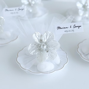 May include: A white wedding favour with a flower-shaped decoration. The flower has a pearl centre and crystal details. The favour sits on a small, scalloped dish with gold trim. A tag reads "Mariam & George 09.21.25".
