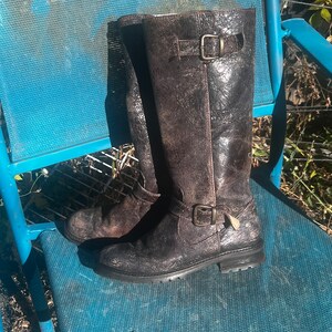 May include: A pair of distressed brown leather knee-high boots with buckle details. The boots have a textured appearance and a worn look, suggesting a vintage or rustic style. The boots are placed on a blue chair.