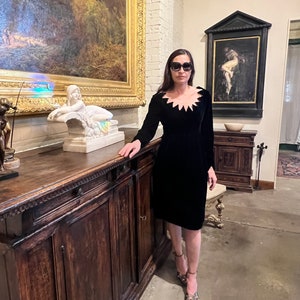 May include: A woman wearing a black velvet dress with a scalloped neckline and black sunglasses stands in a room with a wooden cabinet and paintings on the wall.