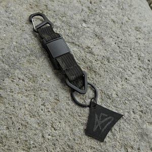 May include: Black keychain with a woven strap, a metal clasp, and a triangular ring. A small, black tag with a white graphic hangs from the ring. The keychain is set against a textured, gray stone surface.