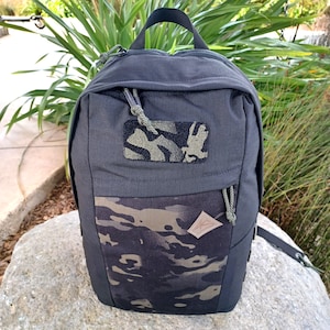 Peut inclure: Un sac à dos noir avec un motif camouflage sur la poche avant et un petit patch camouflage. Le sac à dos a une poignée supérieure, une fermeture éclair et une petite étiquette en cuir. Le sac est posé sur un rocher.