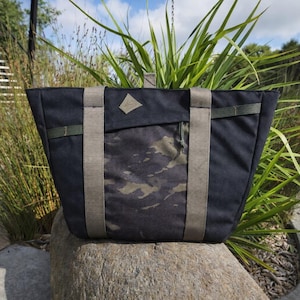 Puede incluir: Bolso tote negro con correas beige y un panel frontal de camuflaje. El bolso tiene un parche en forma de diamante y un pequeño detalle de correa verde. El bolso está sobre una roca.