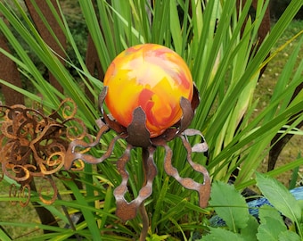 Könnte beinhalten: Dekorative Gartenstele mit einer bunten Glaskugel in Gelb-, Orange- und Rottönen. Die Kugel wird von einer rostigen Metallstruktur mit fließenden Formen gehalten. Grünes Laub bildet den Hintergrund.